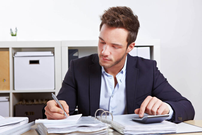 Ein Mann sitzt im Büro an einem Schreibtisch. Vor ihm liegt ein Ordner. Er benutzt einen Taschenrechner und macht sich auf einem Block Notizen.