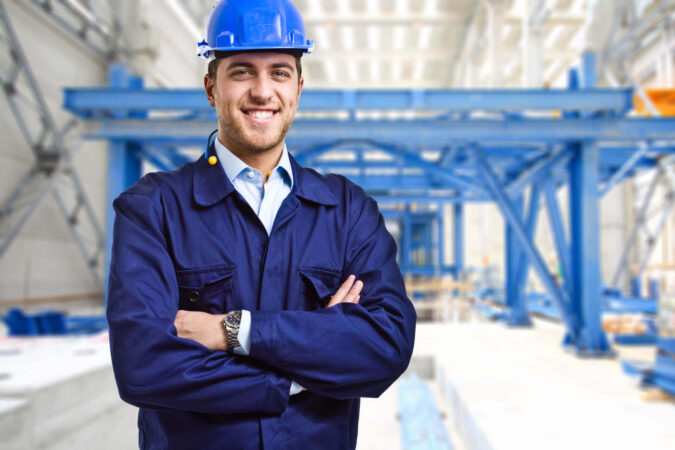Ein Mann mit einem blauen Helm steht lächelnd und mit verschränkten Armen in einer Werkhalle.