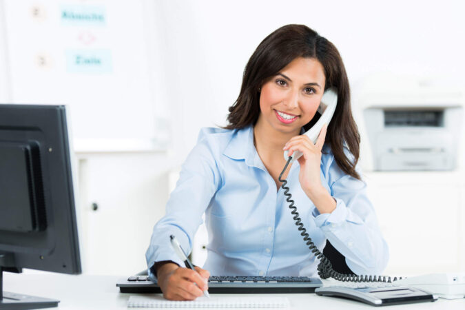Eine braunhaarige Frau sitzt lächelnd an einem Schreibtisch und telefoniert.